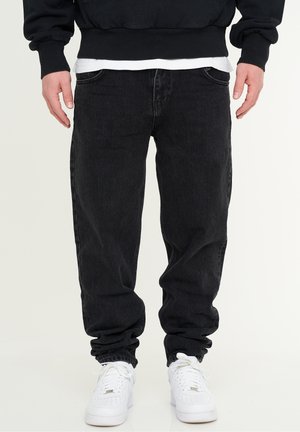 Black denim jeans with a relaxed fit, featuring five pockets and a slightly tapered leg. Worn with white athletic sneakers.