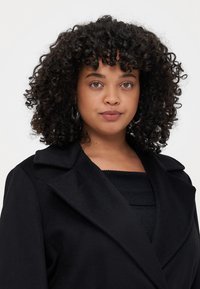Cappotto di lana nero con un ampio colletto, caratterizzato da un design strutturato e una texture liscia; indossato sopra un top nero a coste.
