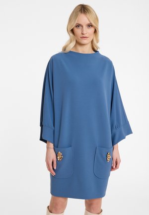 Frau in einem lockeren blauen Kleid mit breiten Ärmeln und zwei vorderen Taschen, verziert mit goldenen abstrakten Broschen, steht vor einem weißen Hintergrund.