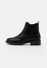 Bota Chelsea de cuero negro con detalles de brogue, paneles laterales elásticos y una etiqueta de agarre. Suela resistente con un leve tacón. Textura suave.