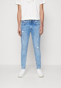 Jeans de mezclilla azul claro con un ajuste slim, que presentan detalles desgastados y un diseño clásico de cinco bolsillos. Combinados con una camiseta blanca.