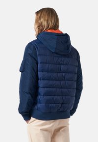 Marinblå pufferjacka med huva, med horisontell quiltning, ribbad mudd och en orange foder, tillverkad av lätt, blankt material.