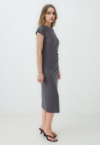 Robe midi grise à manches courtes avec une silhouette ajustée et un détail froncé sur le côté. Associée à des tongs à talons noirs, se tenant sur un fond clair.