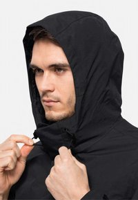 Zwarte waterdichte jas met een aansluitende capuchon, verstelbare trekkoorden en een gladde textuur. Bevat een close-up detail van een hand die de rits vastgrijpt.