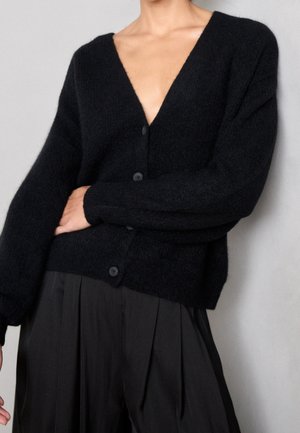 Person wearing a black fuzzy buttoned cardigan with a deep V-neck and loose black pleated pants, standing against a plain background.