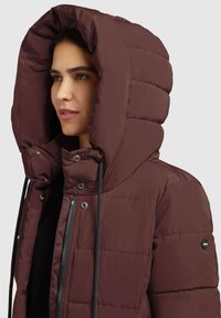 Burgundy puffer-jacka med överdimensionerat huva, quiltad textur, dragkedjor och justerbar snörning. Har tryckknappar och en logopatch.
