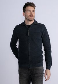 Donkerblauwe zip-up trui met een gestructureerd breipatroon, een neklijn en contrasterende materialen rond de rits. gedragen over een zwart shirt.