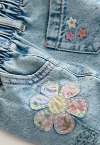 Tessuto di denim con un lavaggio blu chiaro presenta un'applicazione floreale ricamata sulla tasca. Include fiori ricamati in rosa, bianco e giallo.