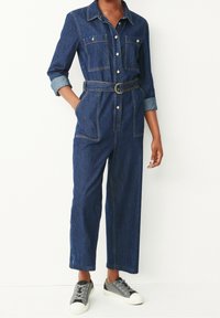 Combinaison en denim à manches longues, avec deux poches sur la poitrine, une taille ceinturée et des poignets retroussés. Elle présente une couleur bleu foncé et des détails de couture visibles.