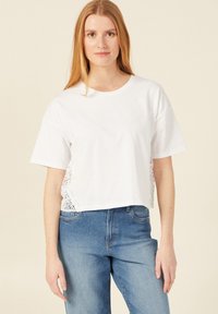 Witte cropped t-shirt met korte mouwen en kantdetails aan de zijkanten, gecombineerd met lichtblauwe high-waisted jeans.