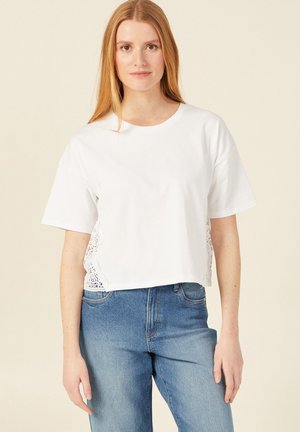 T-shirt blanc court à manches courtes avec des détails en dentelle sur les côtés, associé à un jean taille haute bleu clair.