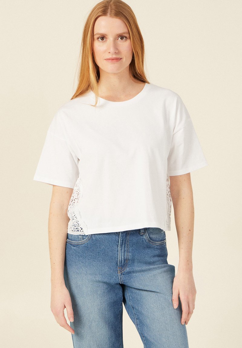 Witte cropped t-shirt met korte mouwen en kantdetails aan de zijkanten, gecombineerd met lichtblauwe high-waisted jeans.