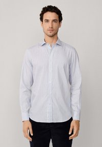 Hombre vestido con una camisa de botones de manga larga a cuadros en azul claro y pantalones oscuros, posando contra un fondo gris liso.