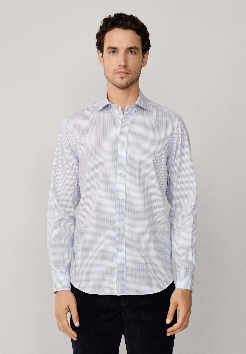 Hombre vestido con una camisa de botones de manga larga a cuadros en azul claro y pantalones oscuros, posando contra un fondo gris liso.