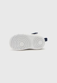 Sola de borracha branca com padrões de trilhos circulares, apresentando uma alça de tecido azul escuro para um ajuste seguro. Sem marcas visíveis na sola.