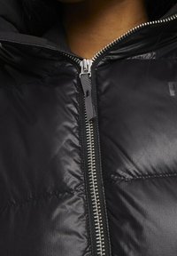 Černá péřovka se zipem vpředu, texturovanou látkou a kapucí. Mezi výrazné detaily patří lesklý zip a mal�é logo.