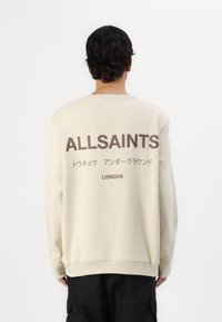 Beige színű, hosszú ujjú pulóver, nagy sötétbarna "ALLSAINTS" logóval és japán karakterekkel a hátsó részén. Egyszínű elülső design.