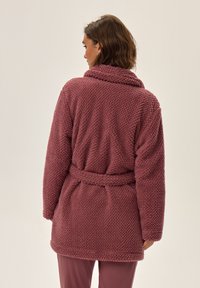 Bordowa szlafrok z polaru o teksturowanym, puszystym wzorze. Posiada kołnierz typu szal, długie rękawy i zapięcie na pasku w talii.