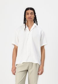 Camicia bianca a maniche corte con un motivo geometrico testurizzato, abbottonatura frontale, colletto a punta, abbinata a pantaloni beige chiaro.