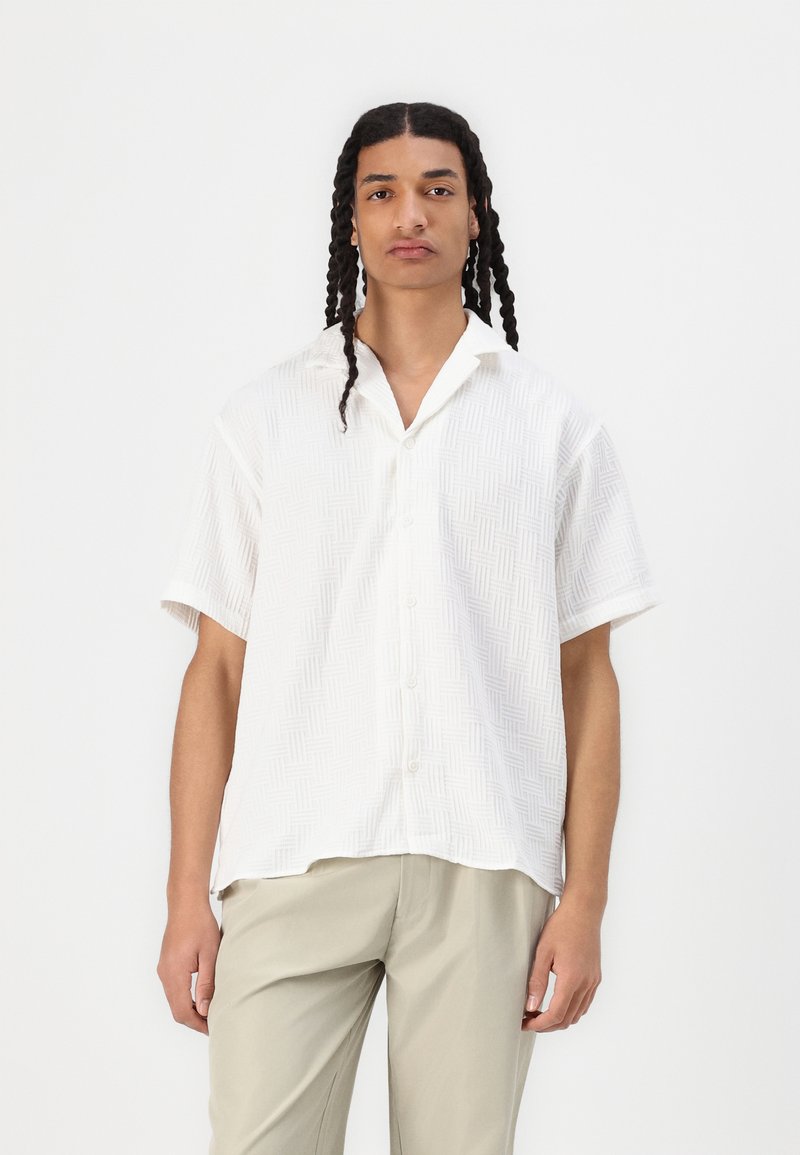 Camicia bianca a maniche corte con un motivo geometrico testurizzato, abbottonatura frontale, colletto a punta, abbinata a pantaloni beige chiaro.