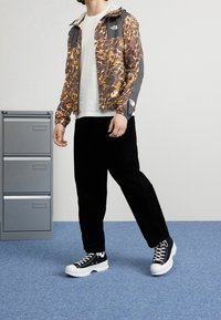 Herrarnas lätta jacka i brun och grå abstrakt mönster, svarta kordfläskbyxor och svarta sneakers med vita sulor.