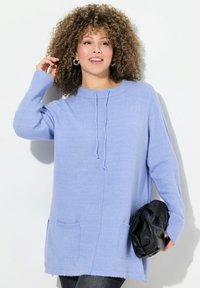 Hellblauer gestrickter Pullover mit langenÄrmeln, hohem Kragen, Kordelzugdetails und zwei vorderen Taschen. Gestylt mit einer schwarzen Clutch.