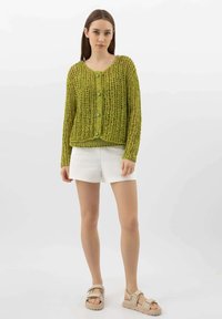 Cardigan verde lavorato a maglia con una vestibilità ampia, motivo testurizzato, scollatura rotonda e cinque grandi bottoni verdi. Abbinato a pantaloni corti bianchi.