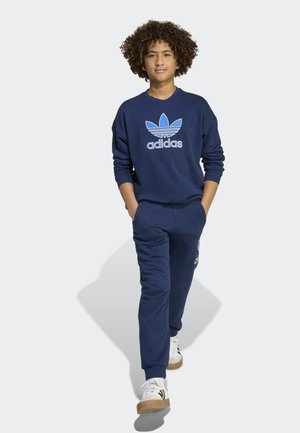 Adolescente con capelli ricci che cammina indossando una felpa Adidas blu navy e pantaloni della tuta abbinati con sneakers bianche.