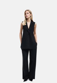 Zwarte mouwloze blazer met een diepe V-hals, gestructureerd ontwerp, twee zijzakken, gecombineerd met brede zwarte broek en puntenhakschoenen.