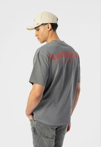 Camiseta gris de gran tamaño con texto rojo en la parte de atrás; combinada con una gorra beige. La camiseta tiene un cuello redondo y mangas cortas, con un ajuste casual.