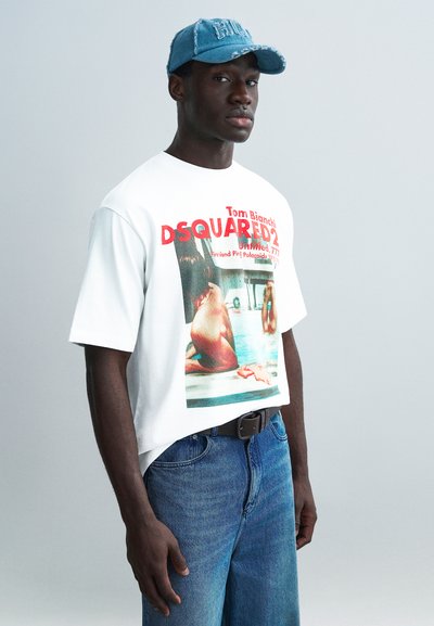 Λευκό υπερμεγέθες T-shirt με φωτογραφικό τύπωμα, κόκκινο κείμενο και κοντά μανίκια. Συνδυασμένο με μπλε τζιν τζιν και μαύρη ζώνη. Ανοιχτό μπλε καπέλο στο κεφάλι.