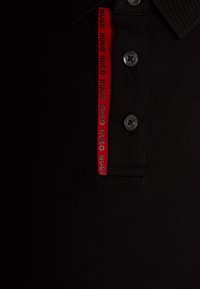 Czarna koszulka polo z teksturowanej tkaniny, dwa czarne guziki oraz czerwony akcent z powtarzającym się logo "HUGO" na kołnierzyku.