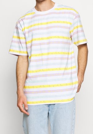 Homme portant un t-shirt rayé pastel avec des lignes horizontales jaunes, violettes et bleu clair, et un jean en denim bleu clair, debout devant un fond blanc.