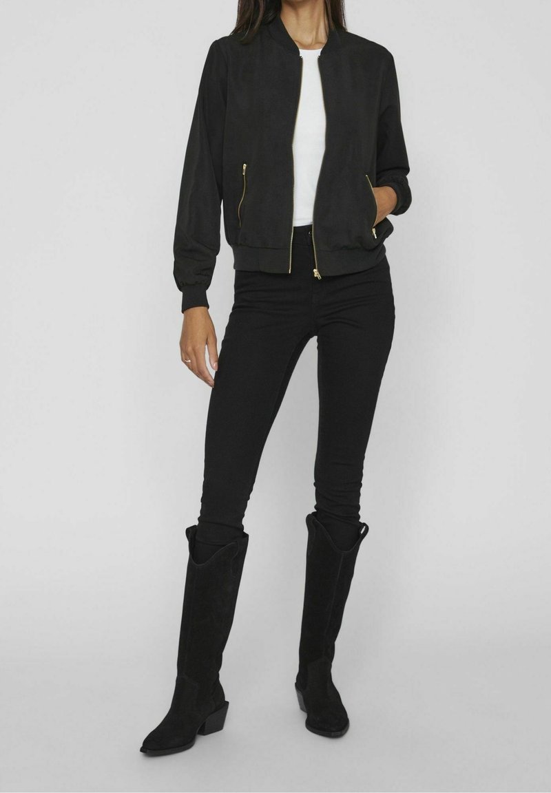 Femme portant une veste bomber noire avec des fermetures éclair dorées, une chemise blanche, un jean skinny noir et des bottes noires montantes, sur un fond uni.