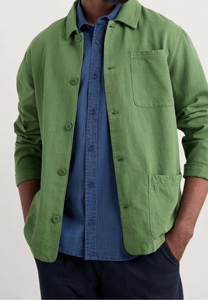 Mann trägt offene grüne Jacke über blau gestreiftem Hemd, die Hände in den Taschen einer dunklen Hose, vor hellem Hintergrund.