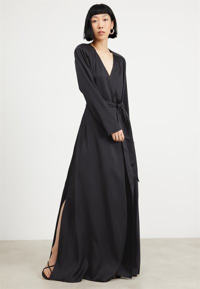 Robe noire à manches longues avec un décolleté en V profond, fente avant et taille nouée, fabriquée dans un tissu lisse. Accentuée avec des sandales à lanières.
