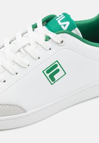 Baltas sneakers ar zaļiem akcentiem, izgatavoti no sintētiskā materiāla. Iespējam teksturēts mēles gabals, zaļš apkakle un logo vidusdaļā.