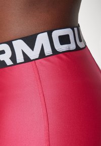 Růžové sportovní legíny mají černý elastický pás s bílým nápisem "Under Armour", hladkou texturu a viditelné detaily prošívání.