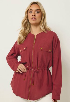 Woman with long blonde hair wearing a long-sleeve red zip-up jacket with front pockets and a drawstring waist, posing against a plain background.