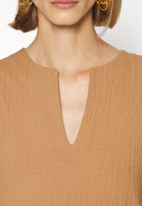 Femme portant un haut texturé couleur camel avec une encolure en encoche et de grandes boucles d'oreilles créoles dorées, visible du cou aux épaules.