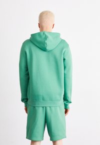 Person med kort blont hår som bär en grön huvtröja och matchande shorts, står vänd bort mot en vit bakgrund.