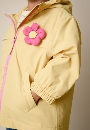 Kind trägt eine hellgelbe Jacke mit rosa Reißverschluss und einer rosa Stoffblume an der Brust, Hand in der Tasche.