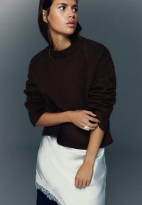 Pulover maro tricotat cu guler înalt, croială largă și mâneci texturate. Potrivit cu o fustă albă din satin cu detalii din dantelă.