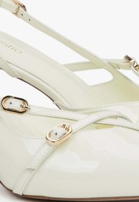 Białe lakierowane buty na słupku z odkrytymi piętami, z delikatnym obcasem, z krzyżującymi się paskami, złotym wykończeniem oraz gładką teksturą.