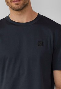 Marineblaue Baumwoll-T-Shirt mit Rundhalsausschnitt, ausgestattet mit einem strukturierten schwarzen Logo-Patch auf der linken Brust. Glatter Stoff mit entspannter Passform.