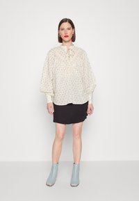 Blusa de creme claro com estampa floral, decote com laço franzido e mangas volumosas, combinada com uma saia preta justa e botas de tornozelo azuis.