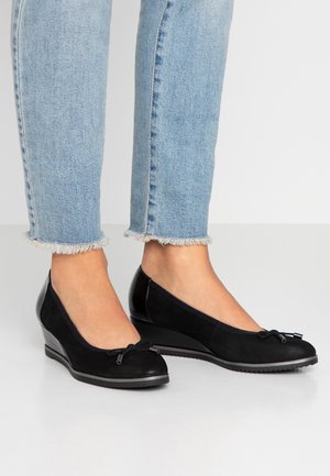 Fødder iført sorte kilehælssko med små sløjfer, kombineret med lyseblå jeans med frynset kant på en hvid gulv.