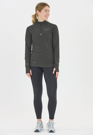 Kvinna i svart långärmad träningströja och leggings, som bär gråa löparskor, stående och ser åt sidan, ler.