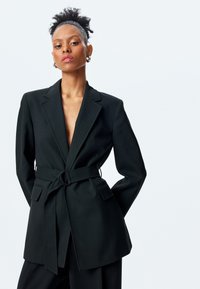 Blazer negro con solapas en pico, cintura ceñida con cinturón y bolsillos frontales. Tejido suave, corte entallado y diseño minimalista.