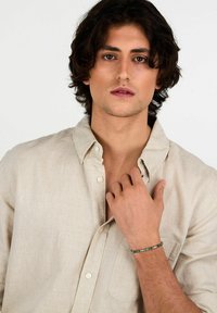 Camicia di lino beige chiaro con bottoni, abbinata a un bracciale di perline con dettagli verdi e trasparenti, indossato su una mano con pelle esposta.